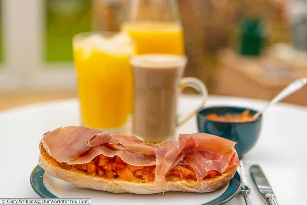 A single slice of the Pan con Tomate, topped with Serano ham on a table in our conservatory. In the background, slightly out of focus, is a carafe of orange juice, a glass of orange juice and a latte coffee in a tall glass cup.