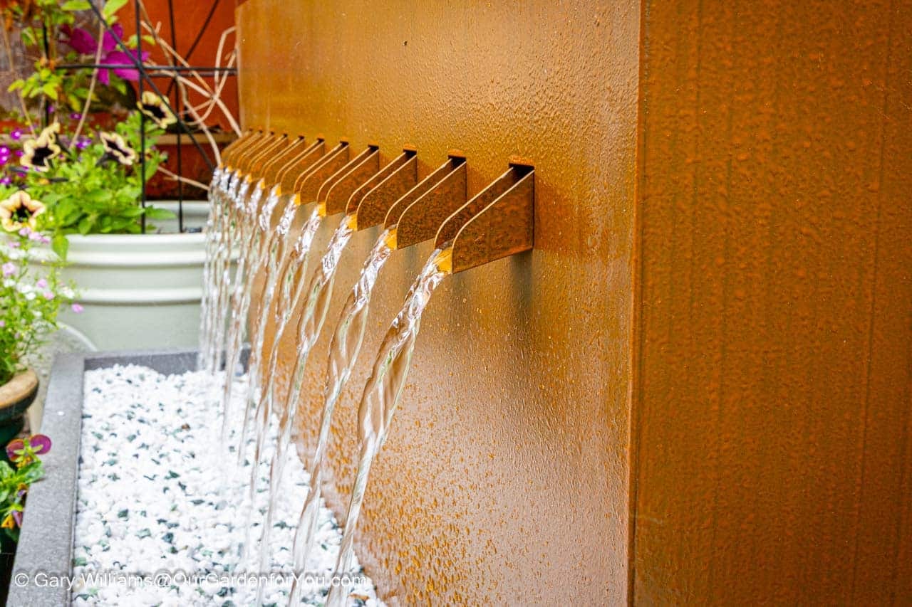 Close-up of our new contemporary water feature, with multiple stainless steel spouts from which clear water cascades onto a bed of white pebbles in our English country garden. The warm, rusted tones of the feature's wall contrast beautifully with the fresh green and colourful flowering plants in the background, adding a tranquil element to the garden.