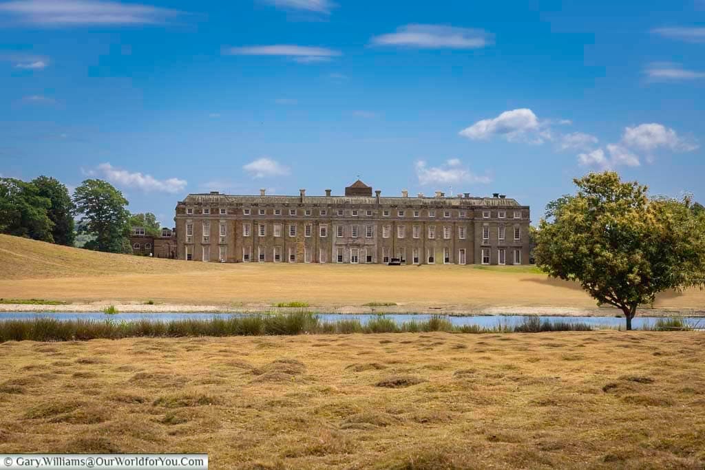A scenic view of the magnificent Petworth House in West Sussex, seen across a serene pond under a bright blue sky. This historic National Trust property is set within its famous Capability Brown landscaped parkland, with golden grasses in the foreground on a sunny summer day.